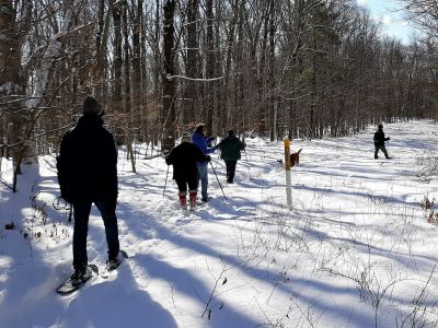 Photograph of a small group of hikers walking on a snow-covered path in the woods of Speer Preserve with snow shoes.