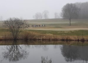 Hikers crossing Wintonbury Hills Golf Course as viewed from across pond with leafless tree silhouette reflected on the water