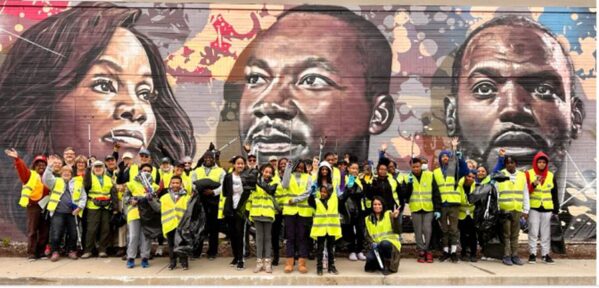 A large group of adult volunteers wearing yellow safety vests stand in front of a large mural with three faces