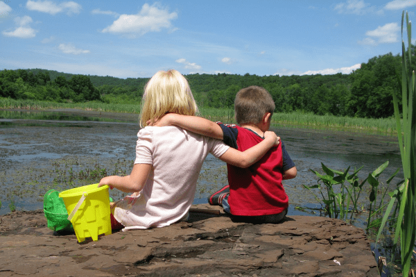 Two children sit on a rock facing away from the photograph with arms around one another's shoulders look out across a pond