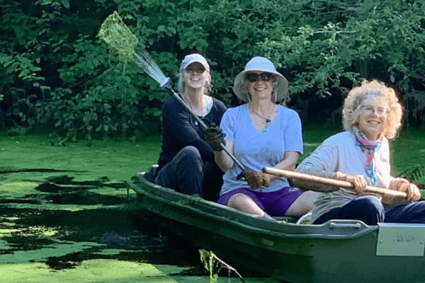 Three volunteers in a canoe facing the photographer with one holding a rake used to remove invasive plants from the water