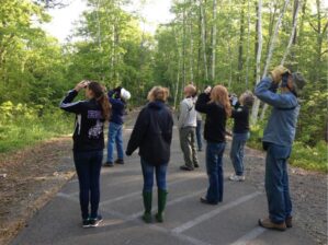 Group of people standing on a paved road look through binoculars toward the trees